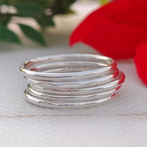 Pu&ograve; includere: Una pila di sei anelli d'argento con un aspetto martellato e strutturato. Gli anelli sono impilati l'uno sull'altro, creando un effetto a strati. Gli anelli sono su uno sfondo sfocato con accenti rossi e verdi.