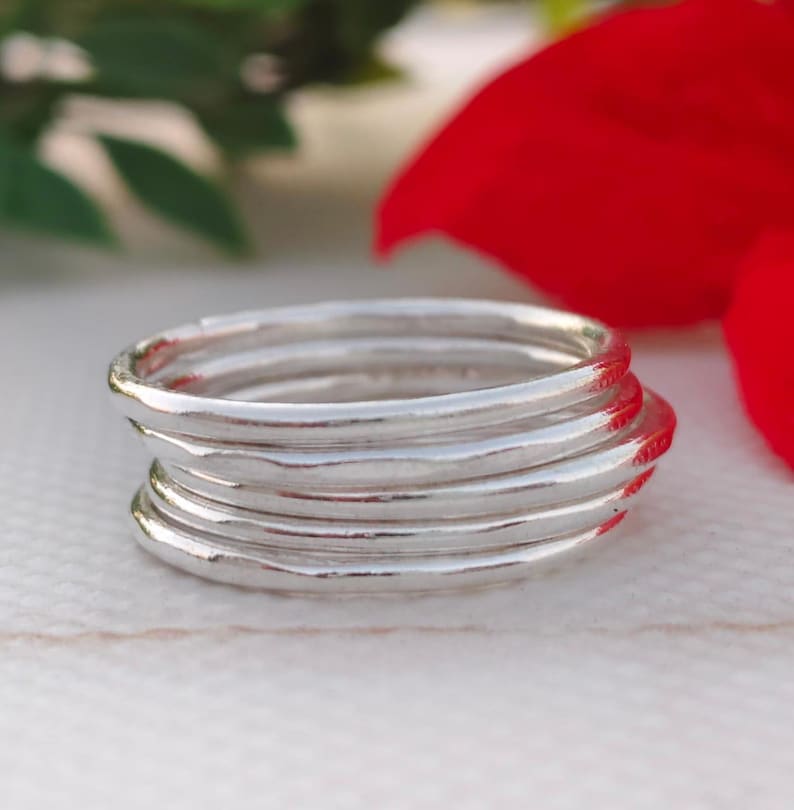 Pu&ograve; includere: Una pila di sei anelli d'argento con un aspetto martellato e strutturato. Gli anelli sono impilati l'uno sull'altro, creando un effetto a strati. Gli anelli sono su uno sfondo sfocato con accenti rossi e verdi.