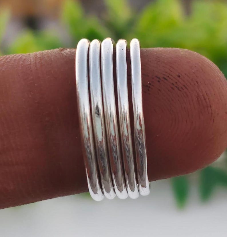 Pu&ograve; includere: Una pila di cinque anelli d'argento, ciascuno con un design liscio e arrotondato. Gli anelli sono mostrati su un dito, con uno sfondo verde sfocato. Gli anelli sembrano essere fatti di metallo lucido.