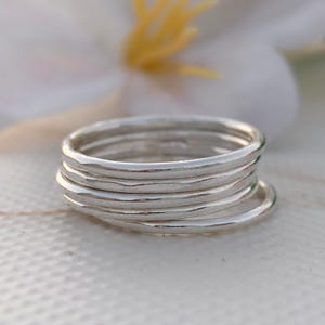 Pu&ograve; includere: Una pila di sette sottili anelli d'argento. Gli anelli sono lisci e lucenti, riflettendo la luce. Gli anelli sono impilati l'uno sull'altro, creando un effetto a strati. Lo sfondo &egrave; sfocato, con una superficie bianca e un fiore.