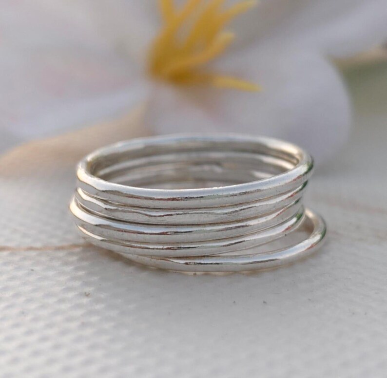Pu&ograve; includere: Una pila di sette sottili anelli d'argento. Gli anelli sono lisci e lucenti, riflettendo la luce. Gli anelli sono impilati l'uno sull'altro, creando un effetto a strati. Lo sfondo &egrave; sfocato, con una superficie bianca e un fiore.