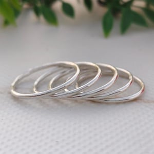 Pu&ograve; includere: Una collezione di sei sottili anelli d'argento disposti in una linea leggermente curva. Gli anelli hanno un design semplice e minimalista, con una finitura liscia e lucida. Gli anelli sono esposti su una superficie bianca, con fogliame verde sullo sfondo.