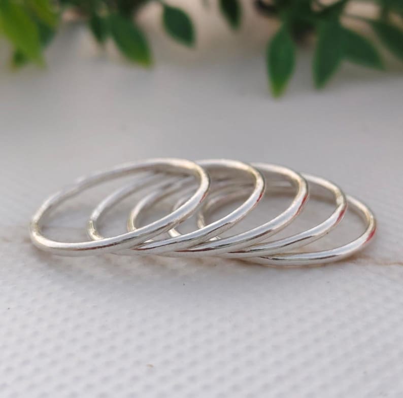 Pu&ograve; includere: Una collezione di sei sottili anelli d'argento disposti in una linea leggermente curva. Gli anelli hanno un design semplice e minimalista, con una finitura liscia e lucida. Gli anelli sono esposti su una superficie bianca, con fogliame verde sullo sfondo.