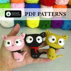 Könnte beinhalten: Drei gehäkelte Katzenfiguren in Rosa, Schwarz und Gelb, jede mit großen weißen Augen und schwarzen Akzenten. Die schwarze Katze hat einen roten Schal. Der Hintergrund umfasst bunte Wolle und den Text "PDF PATTERNS".