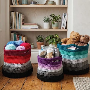 May include: Three handmade crochet storage baskets in red, purple, and teal, each with a black base and gray midsection. The red basket holds yarn, the purple basket holds magazines, and the teal basket contains teddy bears. The baskets are on a wooden floor.