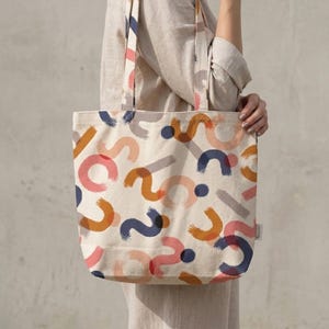 May include: Cream-colored tote bag with a colorful abstract design. The pattern includes orange, pink, blue, and gray brushstrokes and dots. The bag has matching handles and a small tag on the side.