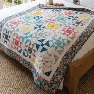 May include: A patchwork quilt featuring geometric patterns in pink, teal, yellow, and navy on a white background. The quilt has a floral border and is displayed on a bed. The quilt is a decorative bedding item.