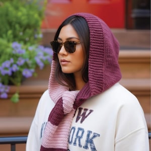 May include: A knitted hood and scarf set in burgundy and light pink. The hood is a solid burgundy colour, while the scarf has alternating stripes of burgundy and light pink. The person is wearing a white sweatshirt with the text "NEW YORK" printed on it.