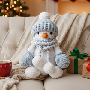Könnte beinhalten: Ein handgefertigter Schneemann-Plüsch mit blauer Strickmütze, Schal und Fäustlingen. Der Schneemann hat eine Karottennase und weiße Bommel. Er sitzt auf einem cremefarbenen Sofa mit einer Decke.