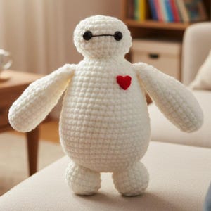 May include: A handmade, white crochet plush toy in the shape of a character with a round body, arms, and legs. The toy has black eyes, a black line for a mouth, and a small red heart on its chest. The toy is sitting on a beige surface.