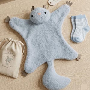 May include: A light blue plush baby blanket shaped like a flying squirrel, with a matching small pair of striped socks and a drawstring bag with the text "Wild Friends Cozy Creature". The blanket has small brown felt feet and ears.