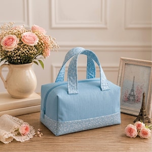 May include: A light blue rectangular fabric bag with two matching handles. The bag features a solid light blue main body and a bottom panel decorated with a small white floral pattern. The handles are also covered in the same white floral print.