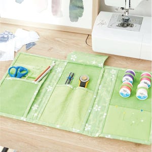 May include: A green fabric sewing organiser with white floral patterns, holding blue scissors, pencils, a rotary cutter, and spools of thread. The organiser is open on a wooden surface, with a sewing machine in the background.