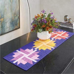 May include: A vibrant, quilted table runner with a geometric starburst pattern in shades of yellow, pink, and orange on a deep blue background. The runner is placed on a dark countertop, with a white vase of flowers and books in the background.