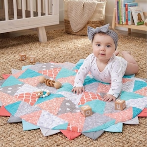 Puede incluir: Un bebé está acostado sobre una colorida colchoneta acolchada. La colchoneta presenta un diseño geométrico con triángulos coral, turquesa y grises. Hay bloques de madera y un sonajero de madera cerca. El bebé lleva un body blanco y una diadema gris.