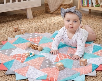 May include: A baby lies on a colorful, quilted play mat. The mat features a geometric design with coral, teal, and gray triangles. Wooden blocks and a wooden rattle are nearby. The baby wears a white onesie and a gray bow headband.