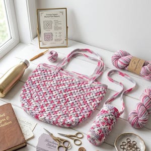 May include: A handmade crochet market tote bag and water bottle holder in shades of pink, gray, and white. The tote bag has two handles. A gold-colored water bottle, yarn, and a crochet hook are also visible.