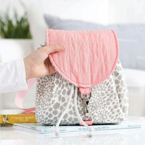 Puede incluir: Una mochila rosa y gris con cierre de solapa. La solapa es de tela rosa acolchada, y el cuerpo principal presenta un estampado de lunares grises y blancos. La mochila tiene un cierre de cordón y un cierre plateado.