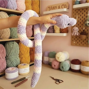 May include: A handmade, plush snake toy in shades of lavender, white, and pink. The snake is made of soft, textured yarn and has small black eyes. The background includes shelves of yarn and the text "Yarn & Co."