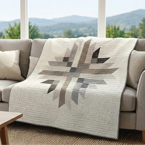Può includere: Una trapunta dai toni neutri con un motivo geometrico a stella drappeggiata su un divano grigio. La trapunta presenta sfumature di grigio e beige, creando un'estetica moderna e minimalista. Lo sfondo mostra una vista panoramica di montagne e alberi.