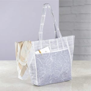 May include: A light gray tote bag with a floral pattern on the front pocket and a striped pattern on the top and handles. A scarf and a small notebook are visible inside the bag. The text "BRAND LOGO" is printed on the top.