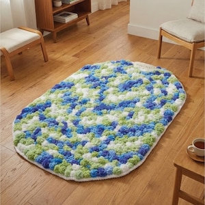 May include: Oval-shaped, handmade rug with a textured, pom-pom-like surface. The rug features a mix of blue, green, and white yarn, creating a colorful, speckled pattern. The rug is placed on a hardwood floor, suggesting a cozy home decor item.