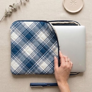 Puede incluir: Una funda acolchada para portátil a cuadros azules y blancos con cierre de cremallera. La funda está abierta, revelando un portátil plateado. Un bolígrafo azul descansa debajo de la funda. Un plato pequeño beige y una ramita de vegetación están en el fondo.