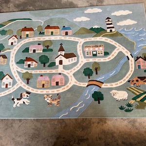 May include: A whimsical rug featuring a town map design with houses, a church, a library, and a lighthouse. The rug includes a road, a river, and illustrations of a dog, a cat, a cow, and a sheep. The text "TOWN LIBRARY" is visible.