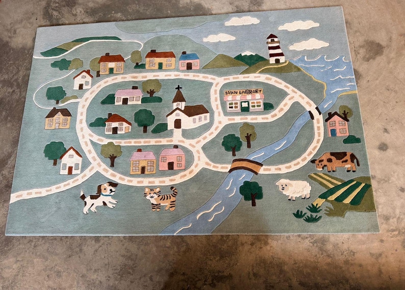 May include: A whimsical rug featuring a town map design with houses, a church, a library, and a lighthouse. The rug includes a road, a river, and illustrations of a dog, a cat, a cow, and a sheep. The text "TOWN LIBRARY" is visible.