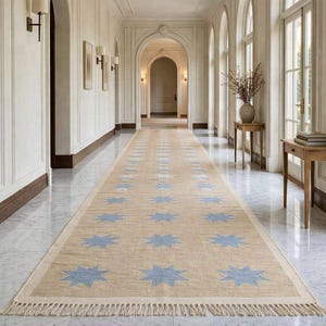 Peut inclure: Un long tapis rectangulaire beige avec un motif répétitif d'éclats d'étoiles bleu clair. Le tapis est placé dans un couloir avec un sol en marbre et des murs blancs. Le tapis a un bord à franges.