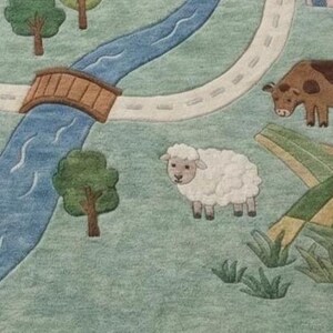 May include: A whimsical, illustrated rug featuring a pastoral scene. A blue river flows under a brown bridge, with a road winding alongside. A sheep and a cow graze on the green grass. Trees and fields complete the charming landscape.