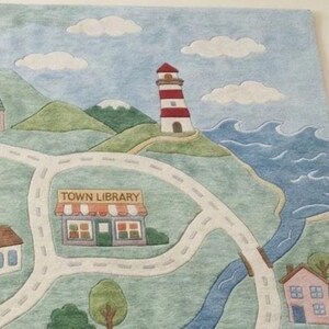 May include: A colorful felt play mat featuring a town map with a lighthouse, houses, roads, a river, and the text "TOWN LIBRARY." The mat has a blue sky with white clouds and green hills.