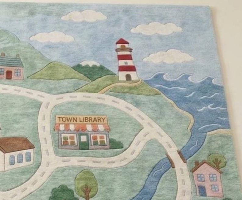 May include: A colorful felt play mat featuring a town map with a lighthouse, houses, roads, a river, and the text "TOWN LIBRARY." The mat has a blue sky with white clouds and green hills.