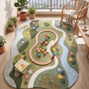 May include: An irregularly shaped area rug featuring a landscape design with shades of green, blue, and white. The rug displays a winding path, a blue river, and small floral motifs. A wooden toy train track is placed on the rug with colorful blocks.