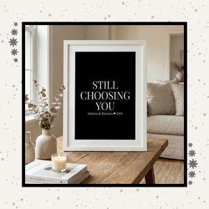 Puede incluir: Un marco blanco que contiene una impresión con el texto "STILL CHOOSING YOU" sobre un fondo negro. El texto incluye los nombres "Sabrina & Mariano" y el año "2010". El marco está sobre una mesa de madera.