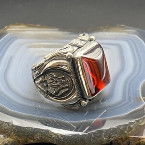 May include: A silver ring with a large, rectangular, red gemstone. The ring has intricate floral and scrollwork designs on the band and around the gemstone setting. The gemstone is a vibrant, transparent red color.