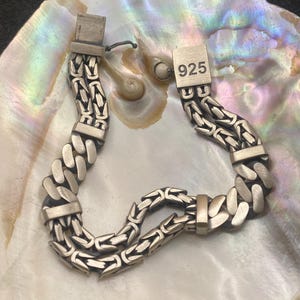 May include: A silver-toned bracelet with a double chain design. The bracelet features a series of interlocking links and rectangular clasps. The clasp is stamped with the number "925". The bracelet is displayed on a pearlescent surface.