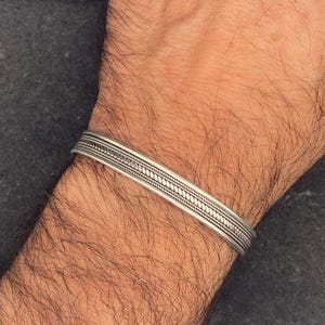 May include: A silver cuff bracelet with a detailed pattern. The bracelet is worn on a wrist with dark hair. The bracelet has a textured design with a series of lines and small dots.