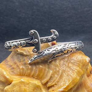 May include: A silver-colored cuff bracelet with an ornate design. The bracelet features a decorative sword-like element at the top and intricate patterns along the band. The bracelet is displayed on a textured, light brown surface.