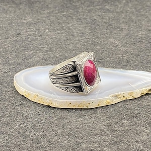 Puede incluir: Un anillo de plata con un diseño ornamentado y una gran piedra preciosa rectangular de color rojo intenso. El anillo está sobre una superficie de piedra blanca y beige sobre un fondo gris. La banda del anillo presenta detalles de volutas.