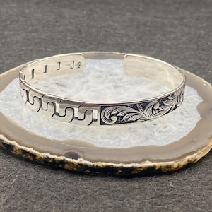 May include: A silver cuff bracelet with intricate engraved designs. One side features a repeating geometric pattern, while the other showcases a detailed floral motif. The bracelet is marked with "925" indicating its sterling silver composition.