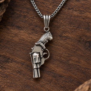 Könnte beinhalten: Ein silberfarbener Metallanhänger in Form eines Revolvers mit texturiertem Griff und zylindrischem Lauf. Der Anhänger hängt an einer silberfarbenen Weizenkette mit polierter Oberfläche und deutlich sichtbaren Metallgliedern.