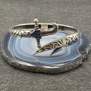 May include: A silver-toned bracelet with an ornate design featuring a dagger motif. The dagger has a black stone-studded hilt and a pointed blade. The bracelet is set on a gray and blue stone surface.