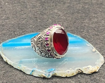May include: A silver ring with an oval, deep red gemstone as the centerpiece. The ring is adorned with smaller, pink gemstones around the main stone and features an ornate, engraved design on the band. The ring is displayed on a blue and white stone.