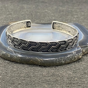 May include: A silver cuff bracelet with a geometric pattern. The bracelet has a textured design and an open back. The cuff is displayed on a gray stone surface. The bracelet is approximately 0.5 inches wide.