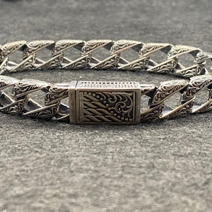 May include: Silver-toned bracelet with an ornate, geometric design. The bracelet features a series of interconnected links with detailed patterns and a rectangular clasp. The metal has a slightly aged appearance, enhancing the intricate details.