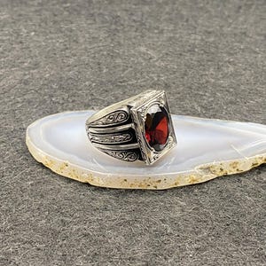 Könnte beinhalten: Ein silberner Ring mit einem ovalen, tiefroten Edelstein, der in einem rechteckigen Rahmen gefasst ist. Der Ring weist auf dem Band und um den Edelstein herum aufwendige Schnörkelarbeiten auf. Der Ring liegt auf einem weißen und beigen Stein.