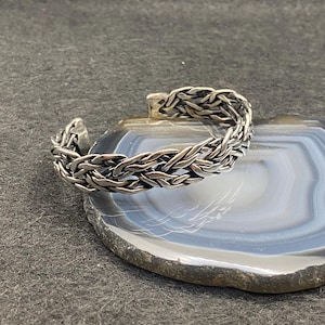 May include: A silver-colored cuff bracelet with a woven design. The bracelet is open at one end and rests on a gray and blue stone surface. The woven pattern gives the bracelet a textured appearance.