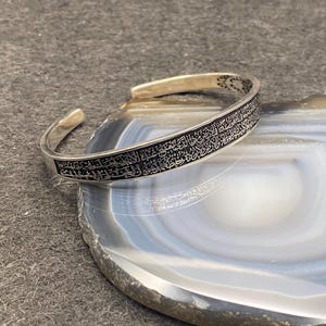 Peut inclure: Un bracelet manchette en argent avec une inscription arabe gravée complexe. Le bracelet ouvert est présenté sur une pierre d'agate grise et blanche, mettant en valeur le design et le savoir-faire de la bijouterie.