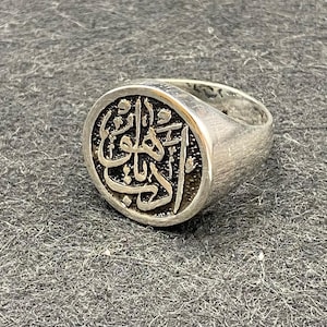 May include: A silver ring with an oval face featuring ornate Arabic calligraphy. The ring has a brushed metal finish and a thick band. The calligraphy is in a darker tone, creating contrast against the silver.